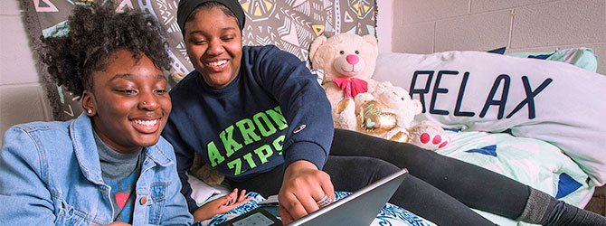 Two students on a bed in a dormatory laughing at someting on a laptop