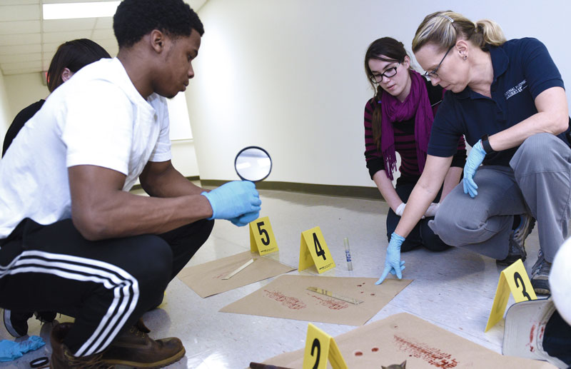 Students in a forensics lab observe as an instructor makes a point about a fictitious crime scene.