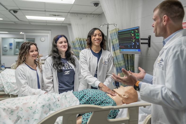 Four people in lab coats stand around a hospital bed with a medical mannequin, while one person demonstrates a procedure and others observe and smile. Medical monitors are visible in the background.