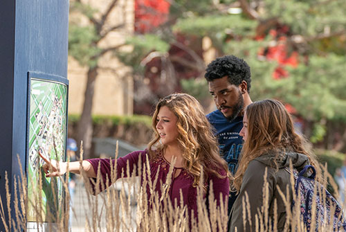 Three college students gather around an outdoor campus map kiosk, with one student pointing to a location, on the University of Akron campus in autumn.