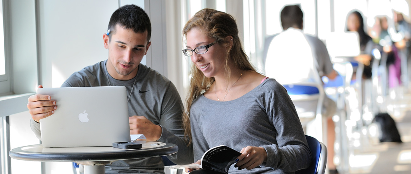 Two college students sit together at a small round table in a bright campus common area, looking at an open laptop. The female student smiles and holds a printed booklet, while the male student points at the screen. Other students are visible in the background.