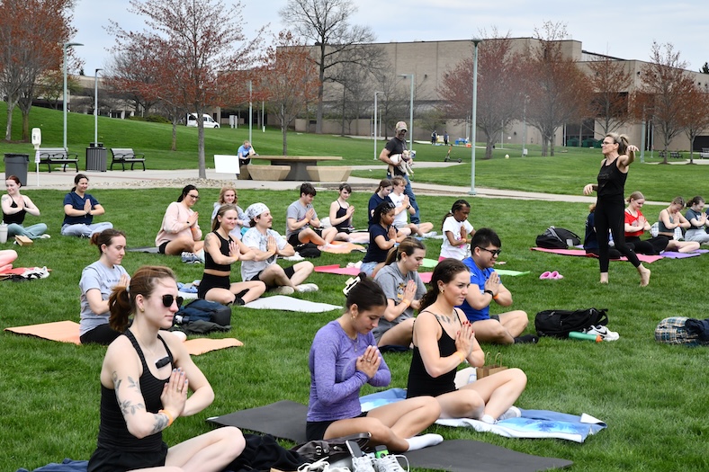 A group of people practice yoga on a grassy lawn, sitting on mats in meditation poses, while an instructor stands in front giving directions. Trees and buildings can be seen in the background.