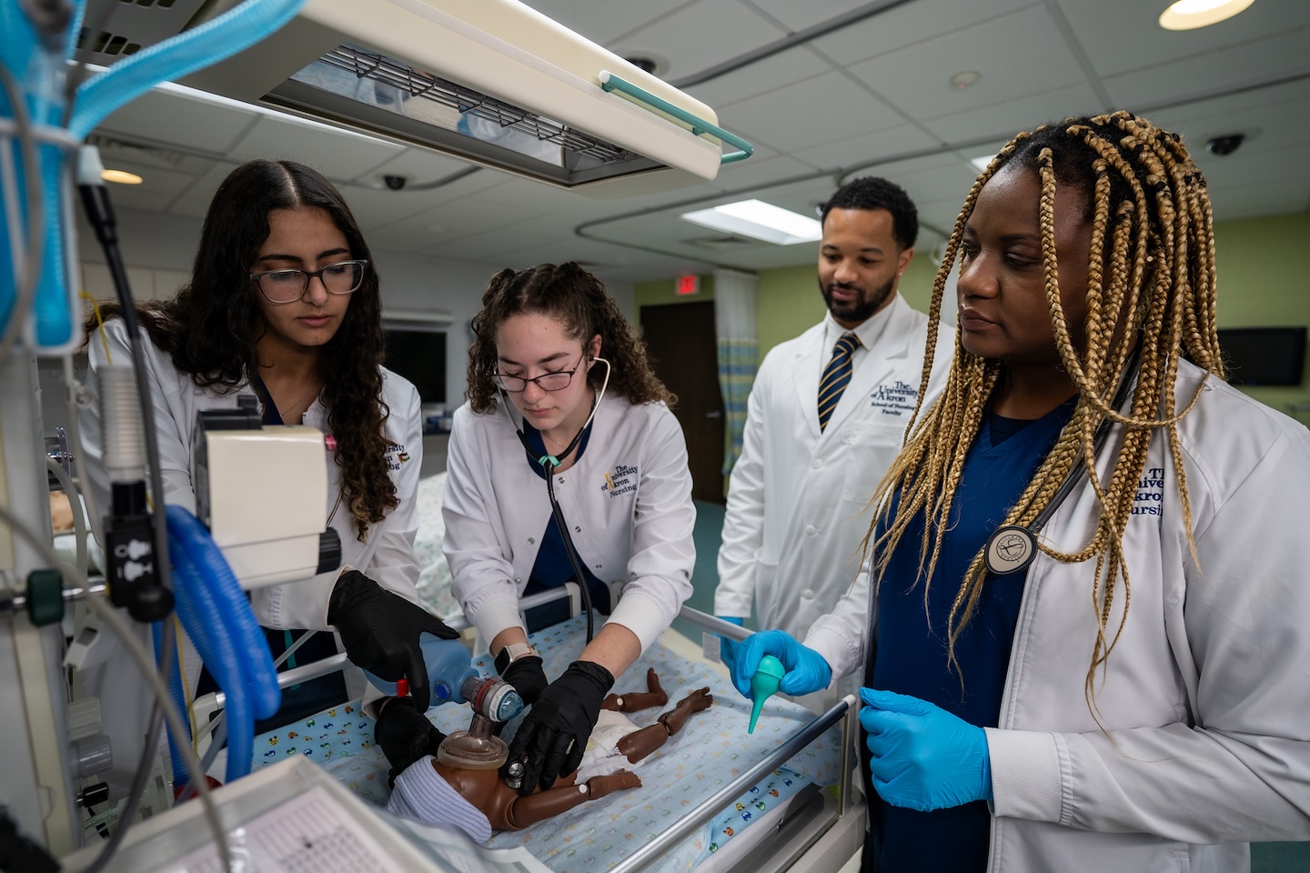 Four nursing students in lab coats practice infant care and resuscitation on a mannequin in a hospital simulation room, using medical equipment and observing closely.