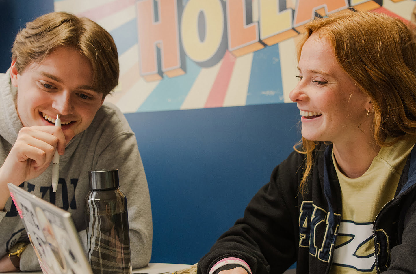 A smiling young man in a grey hoodie holding a pen and a laughing young woman with red hair are sitting together at a table looking at a laptop.