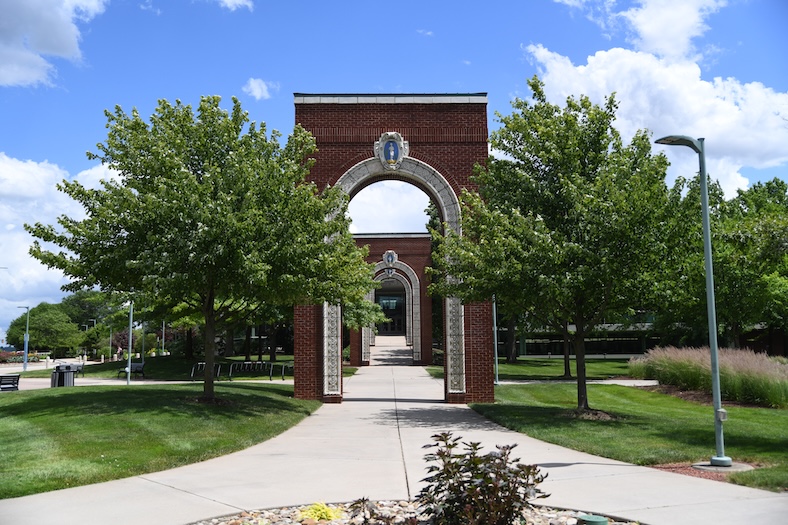 A brick archway stands over a sidewalk surrounded by green trees and neatly trimmed grass on a sunny day with a partly cloudy sky. Another similar arch and a building are visible in the background.