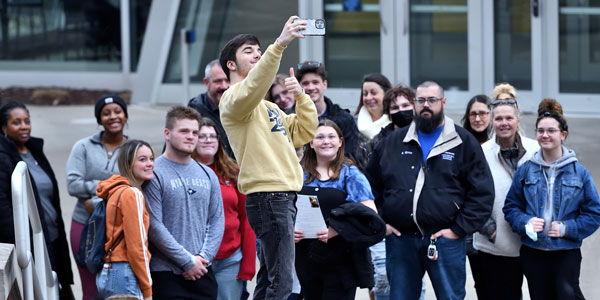 A student in a gold sweatshirt takes a group selfie with a large crowd of prospective students and families gathered outside a university building during a campus tour.