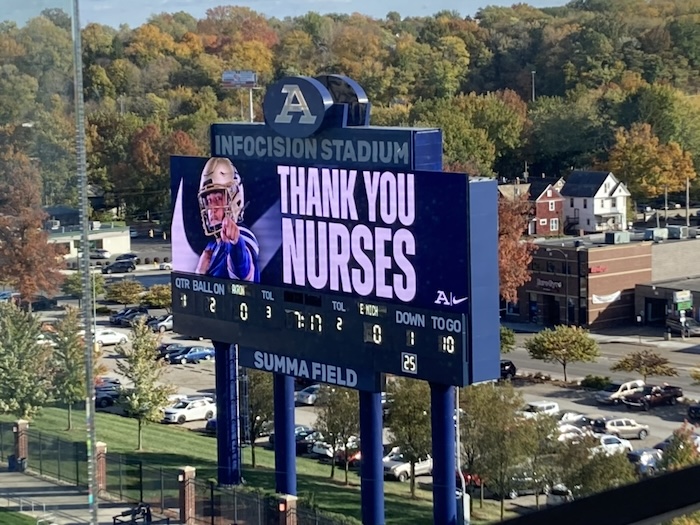 A stadium scoreboard displays an image of a nurse and the message THANK YOU NURSES above the football game information, with trees and parking lot visible in the background.