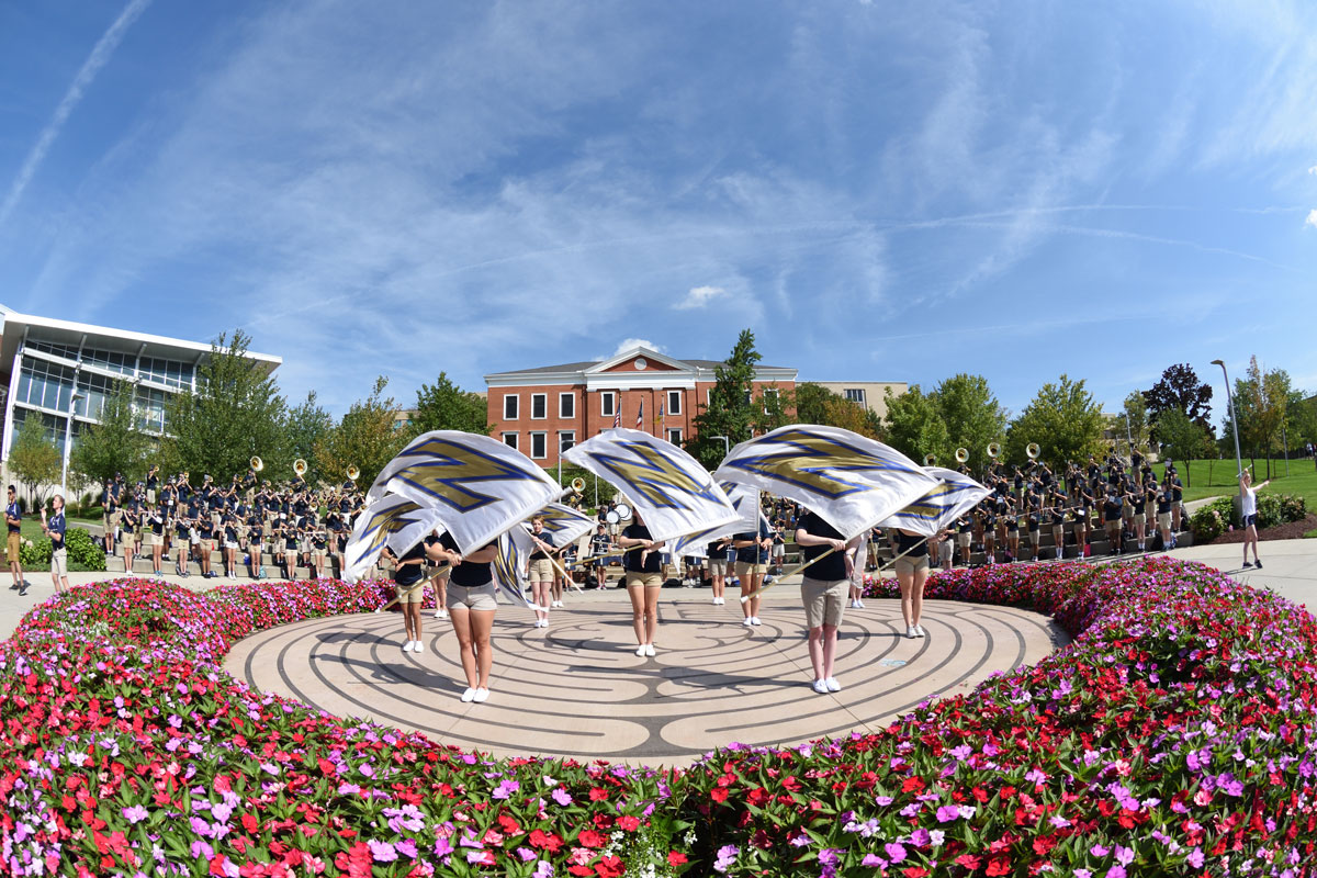 The University of Akron marching band and color guard perform on a circular plaza surrounded by colorful flower beds on a sunny day.
