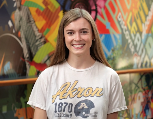 A young woman with long light brown hair smiles at the camera. She is wearing a white “Akron 1870” t-shirt. The colorful, mural-painted wall in the background adds a vibrant and energetic atmosphere.