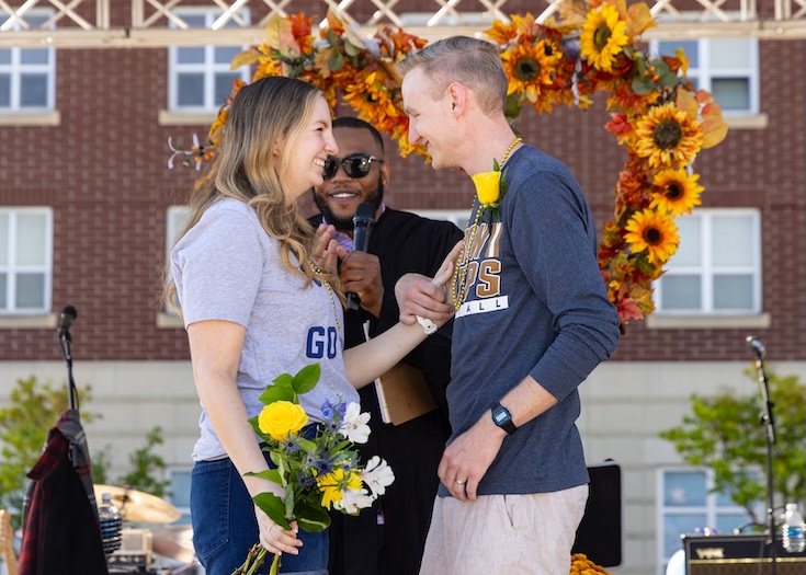 A smiling couple stands onstage with flowers, facing each other, while a person with a microphone speaks behind them. Sunflower decorations and a brick building appear in the background.