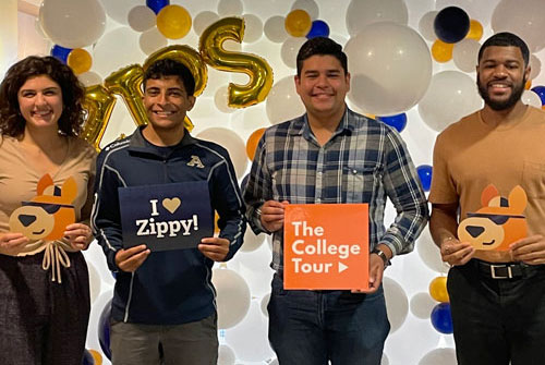 Four University of Akron students smile in front of a balloon backdrop, holding signs reading 'I Love Zippy!' and 'The College Tour,' along with Zippy the kangaroo mascot cutouts.