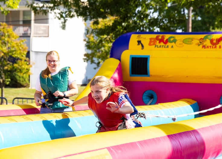 Two people wearing harnesses race on a colorful inflatable bungee run outdoors. One person is leaning forward, stretching, while the other smiles in the background. Trees and a building are visible behind them.