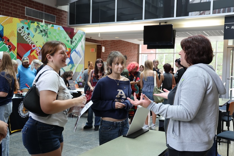 Three people stand and talk near a table with a laptop in a busy indoor area; others are gathered around various tables in the background, and colorful posters decorate the walls.