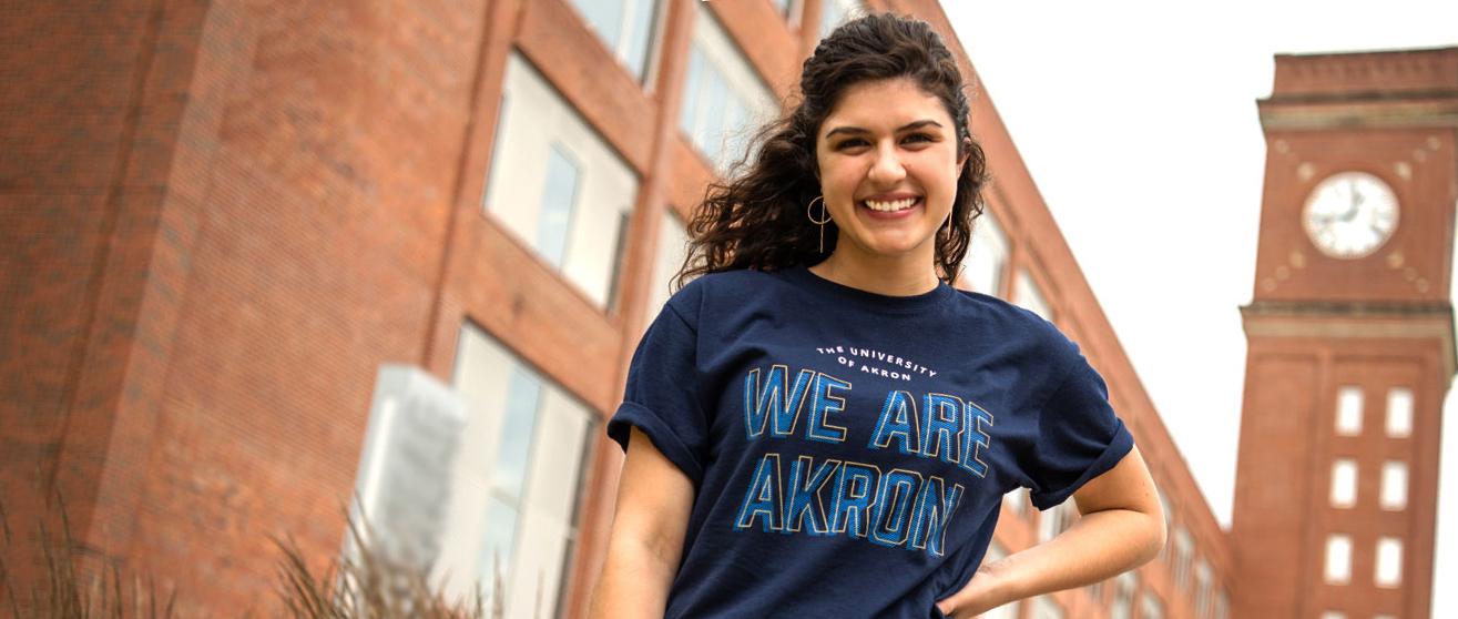 A smiling University of Akron student stands outdoors on campus wearing a navy 'We Are Akron' t-shirt, with brick university buildings and a clock tower visible in the background.
