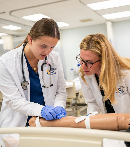 Nursing student practices inserting an IV into a medical simulation arm while an instructor observes and provides guidance.