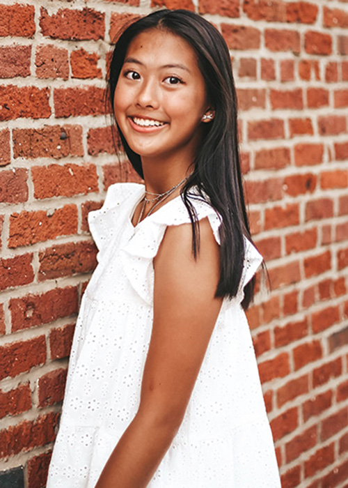Headshot of girl in white dress against brick wall.